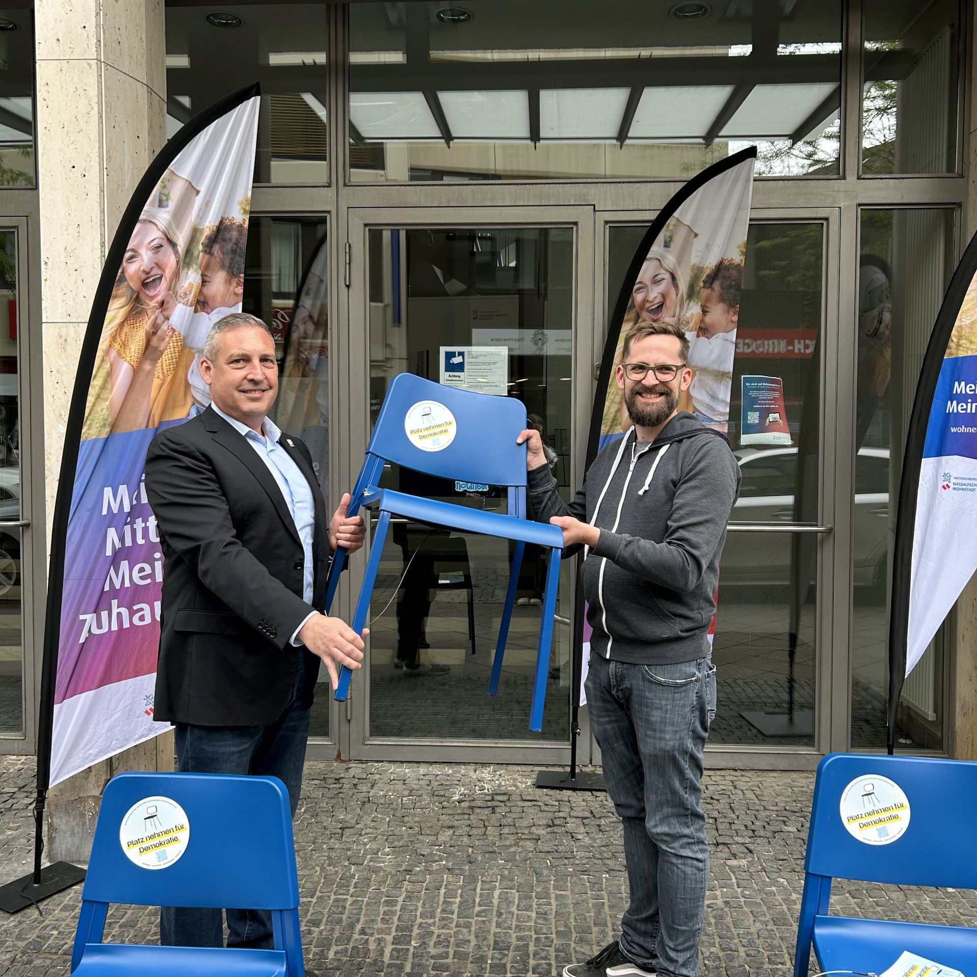 Zwei Männer halten einen blauen Stuhl, im Vordergrund zwei weitere blaue Stühle