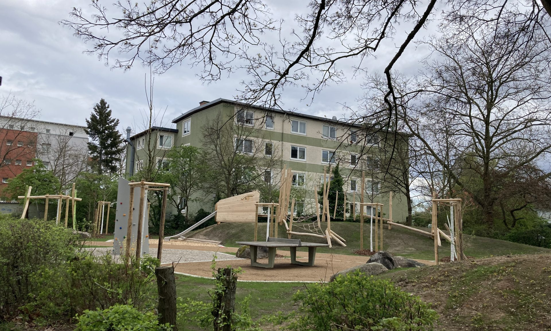 Spielplatz u.a. mit Schaukel, Kletterwand, TT-Platte und Mehrfamilienhäusern im Hintergrund