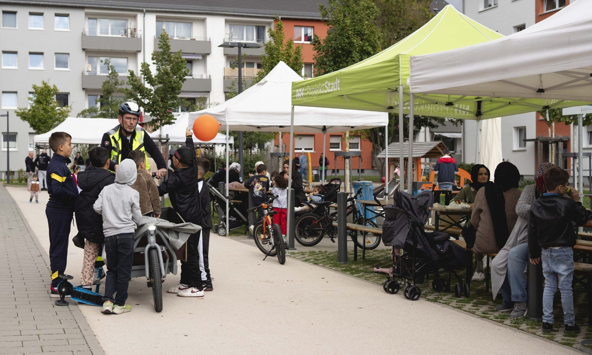Ein Mann auf einem E-Lastenrad, umringt von Kindern, eines davon mit einem Luftballon, an der Seite Menschen, die an Tischen unter Pavillons sitzen.