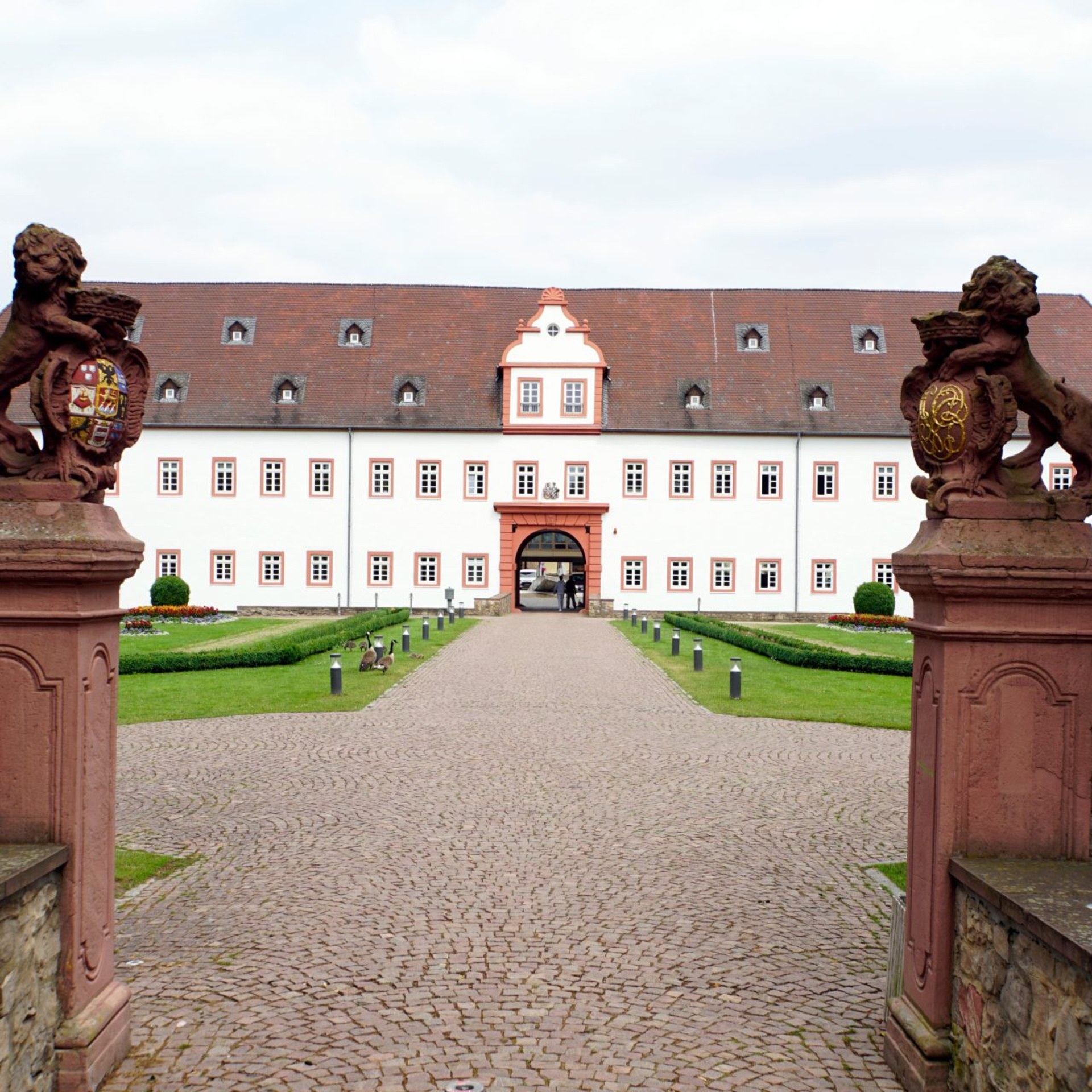 Blick auf ein schlossähnliches Gebäude mit einer gekiesten Auffahrt und zwei steinernen Figuren auf zwei Sockeln im Vordergrund