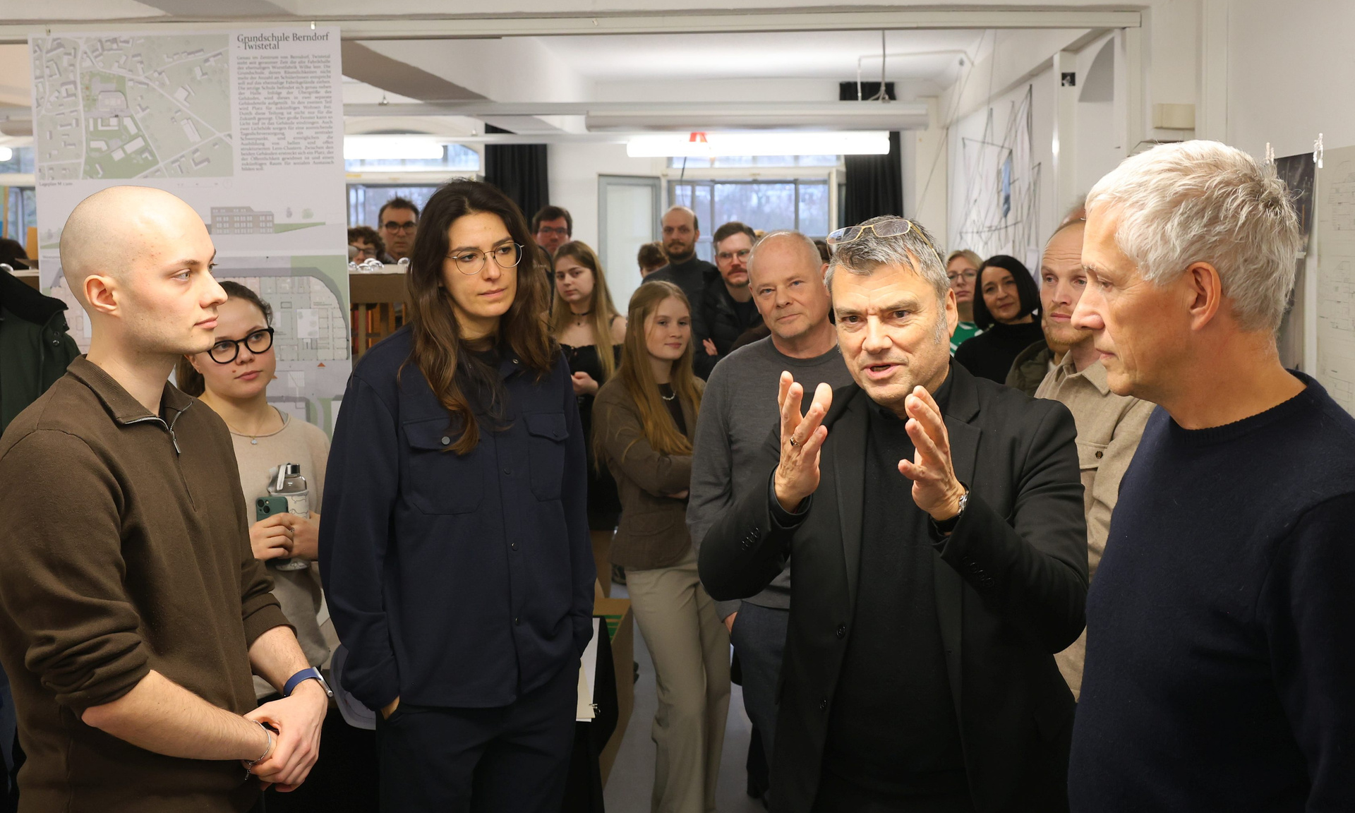 Mehrere Menschen stehen in einer Gruppe beisammen, einer spricht uns gestikuliert, die anderen hören zu, im Hintergrund sieht man Teile einer Ausstellung von studentischen Arbeiten 