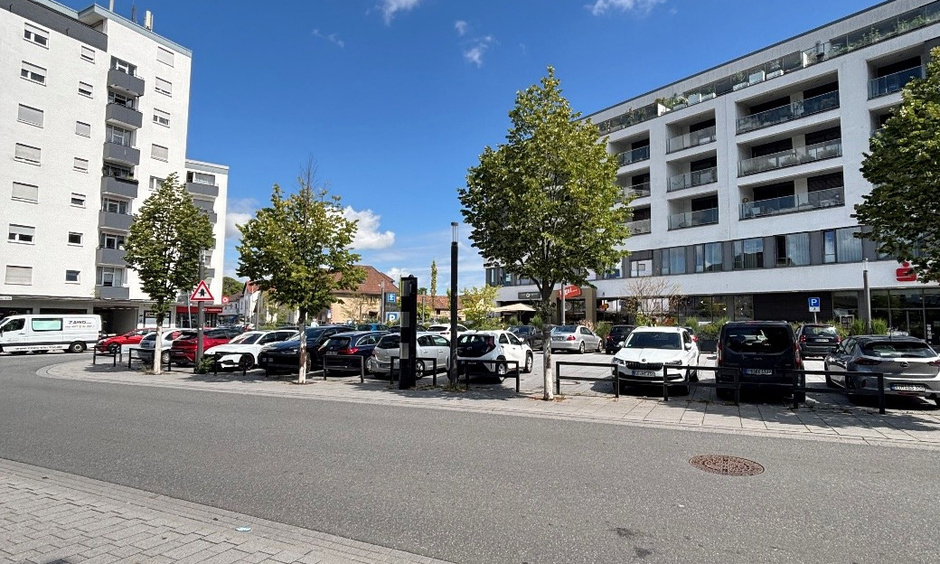Zu sehen ist ein moderner Parkplatz zwischen zwei Wohnhäusern mit Bäumen.
