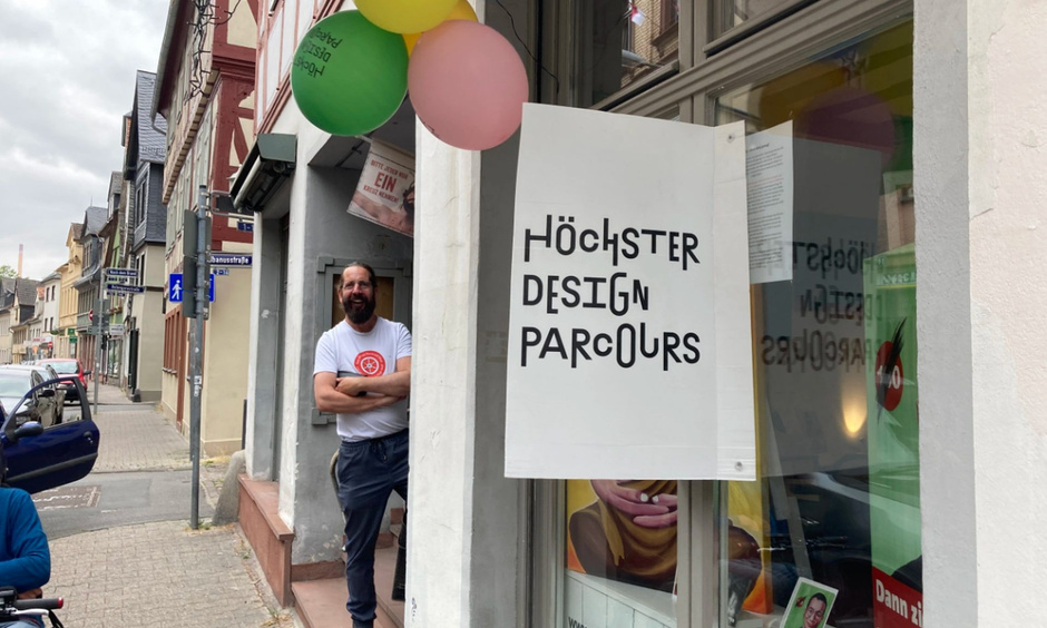 Geschäft, dessen Eingang mit Luftballons geschmückt ist. Ein Mann mit Vollbart steht auf der Türschwelle und schaut Richtung Kamera. Schild mit der Aufschrift "Höchster Designparcours"