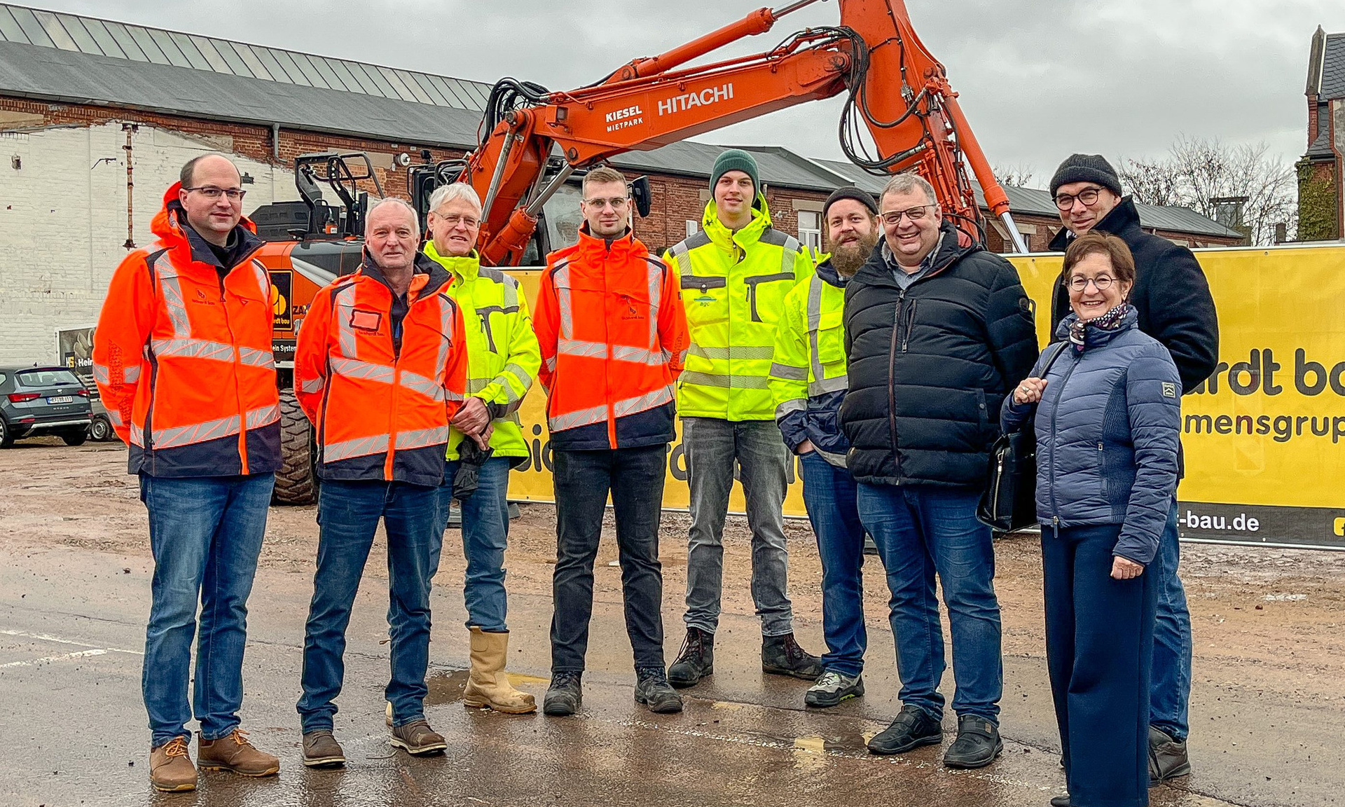 Gruppenfoto zum Baubeginn mit Bad Hersfelds Bürgermeisterin Anke Hofmann (re.), Jörg Albin (2. v. re.), Projektleiter der ProjektStadt, sowie sieben weiteren Personen. Im Hintergrund ein Bagger. Foto: Kreisstadt Bad Hersfeld