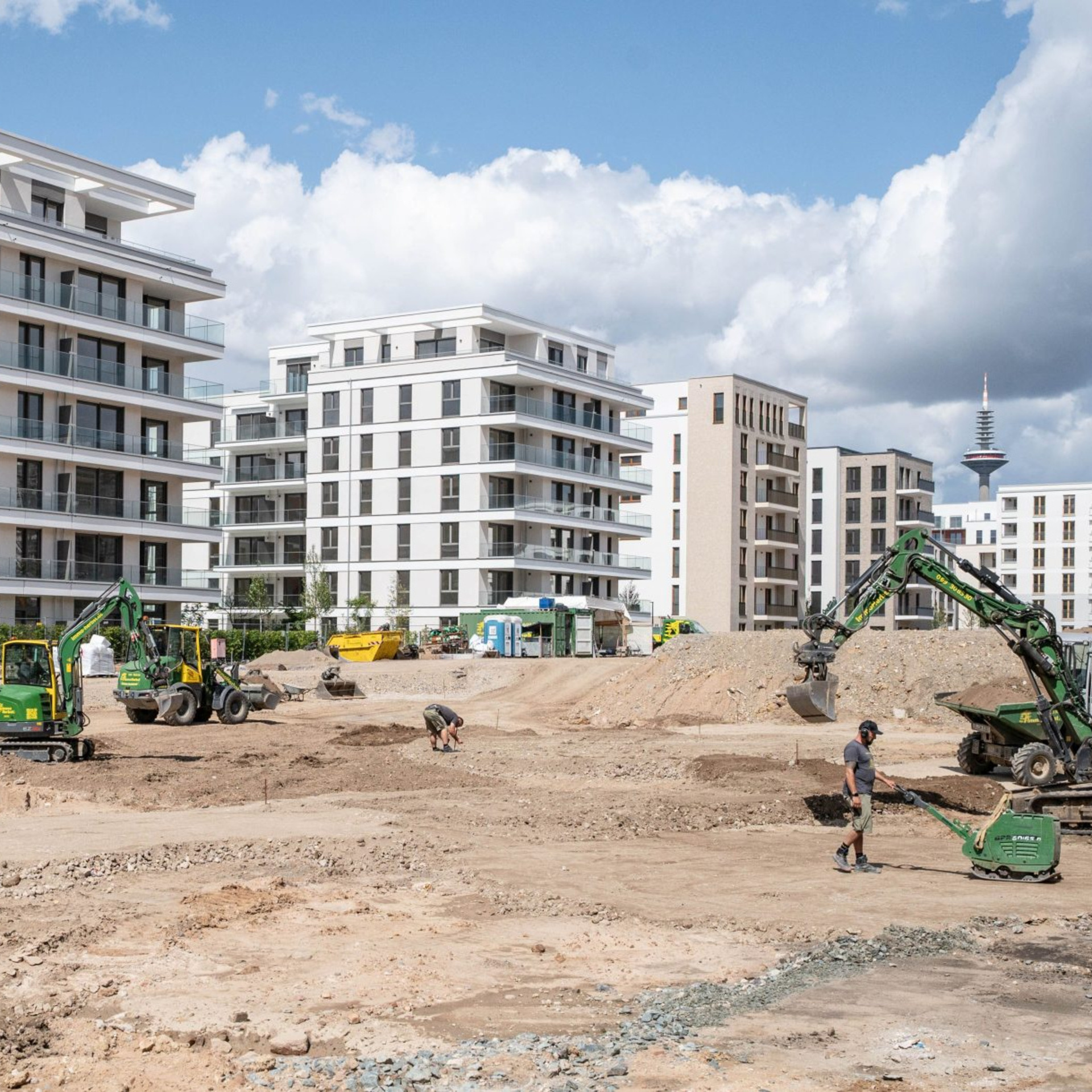 Eine Baustelle mit weitgehend fertiggestellten Neubauten im Hintergrund, im Vordergrund eine riesige Baugrube, Baustellenfahrzeuge und Bauarbeiter.